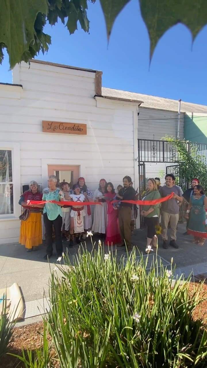 Opening day: a crowd of familia and friends, elders and kids among them, holding a red ribbon in front of the white shop with its carved La Ciendita sign.