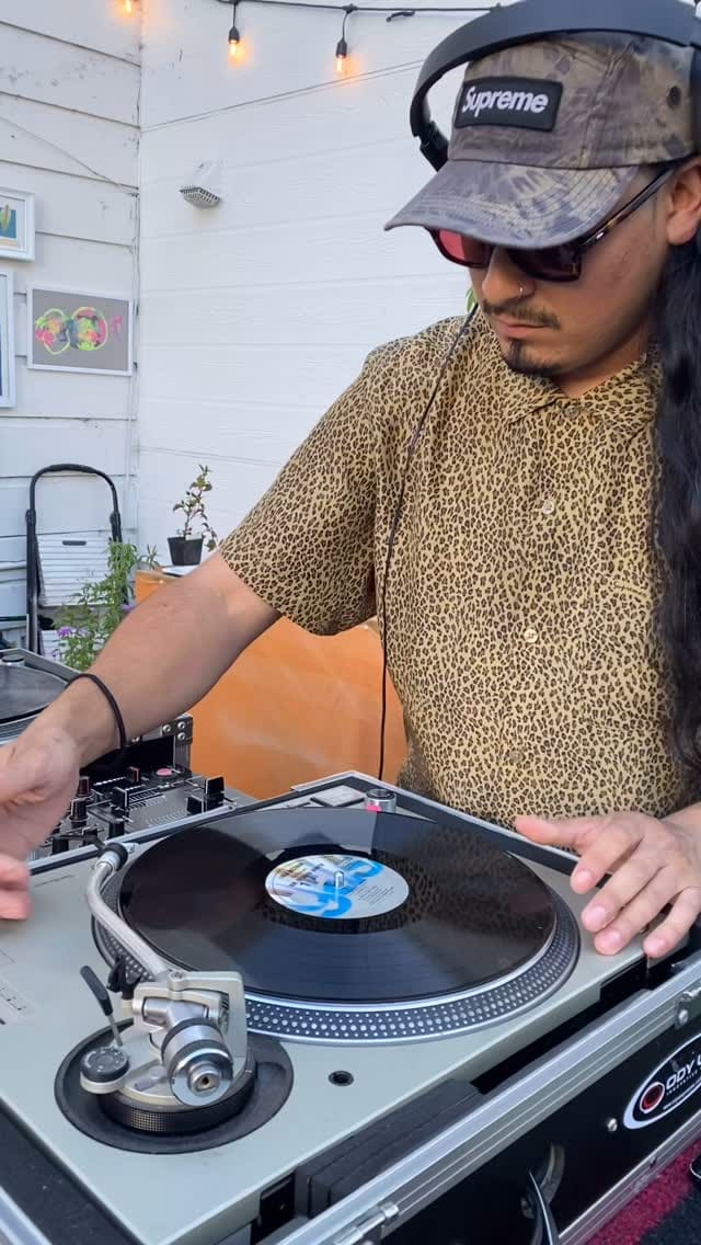 A DJ spinning records in the courtyard at an art pop-up, string lights and plants behind.