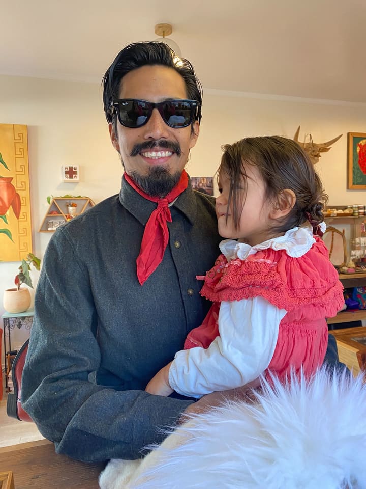 Octavio in sunglasses and a red neckerchief, holding his daughter in a red ruffled dress inside the shop.