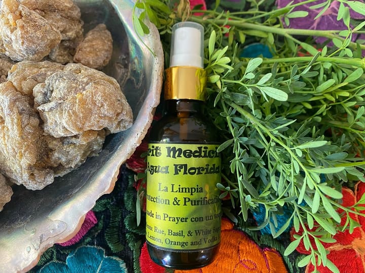 A bottle of agua florida, chunks of copal resin in an abalone shell and fresh ruda, laid on black cloth embroidered with flowers.