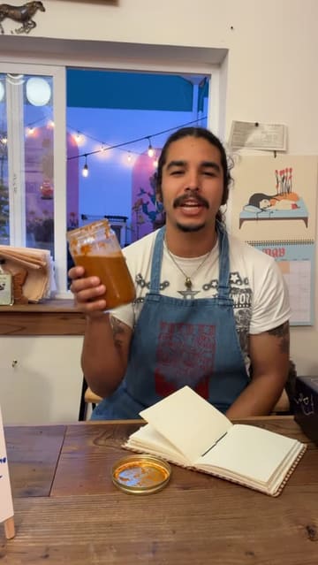 A man in a denim apron holds up a jar of salsa at a community dinner, the lit courtyard and pink arches glowing through the window behind him.