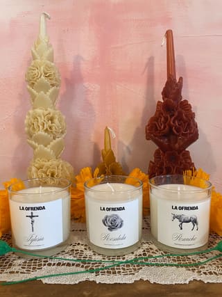 Tall ofrenda candles and carved wax flower candles with paper marigolds, in front of the shop’s pink wall.