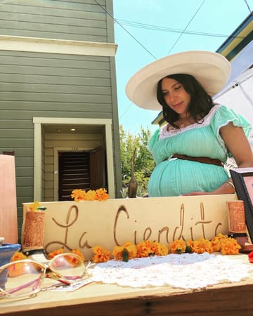 2022: the founder, pregnant, in a wide white hat behind a hand-lettered La Ciendita sign lined with marigolds at the Blue Lake summer market.