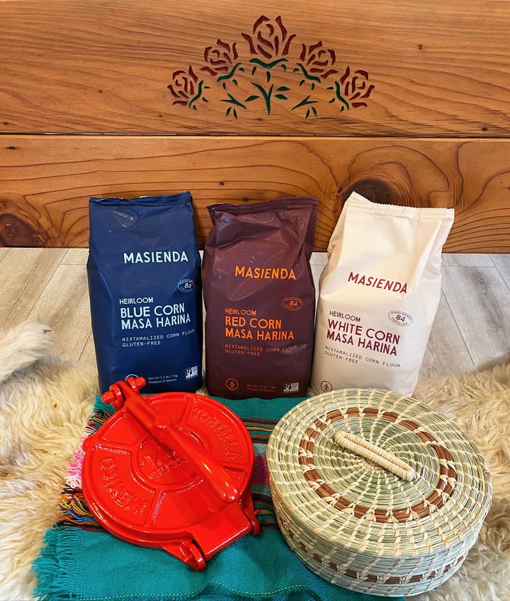 Bags of blue, red and white corn masa harina beside a cast iron tortilla press and a woven tortilla basket.
