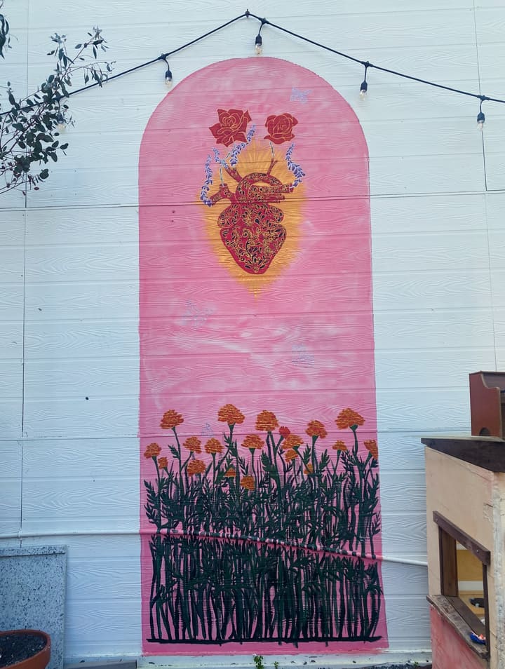 A pink arch mural on the courtyard wall: a red sacred heart with roses and lavender above a field of painted marigolds, under string lights.