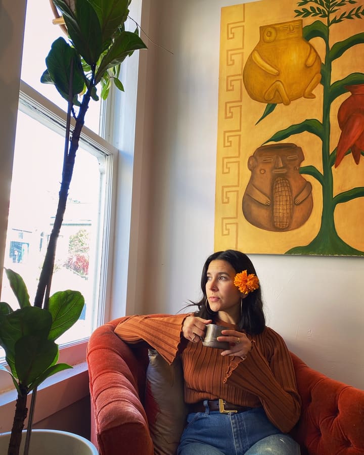 A woman with a marigold tucked in her hair sits in the rust velvet chair holding a mug, under the painting of corn and clay vessels.