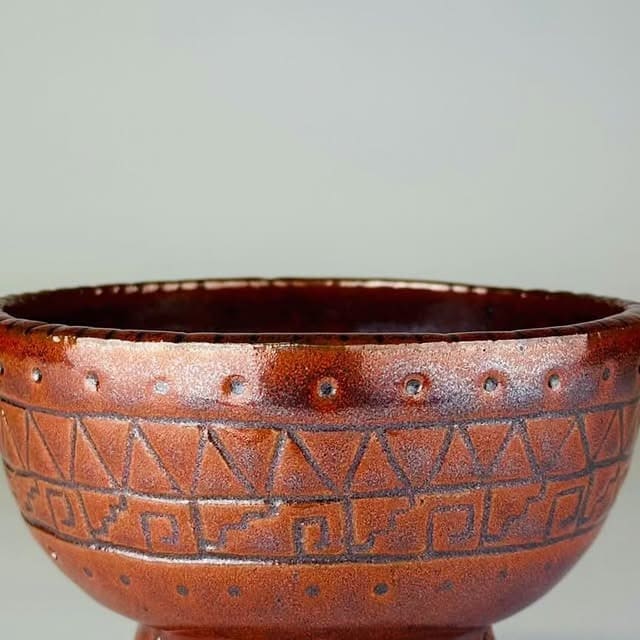 A close view of a reddish brown stoneware bowl with carved triangles and a stepped line, on a plain pale background.