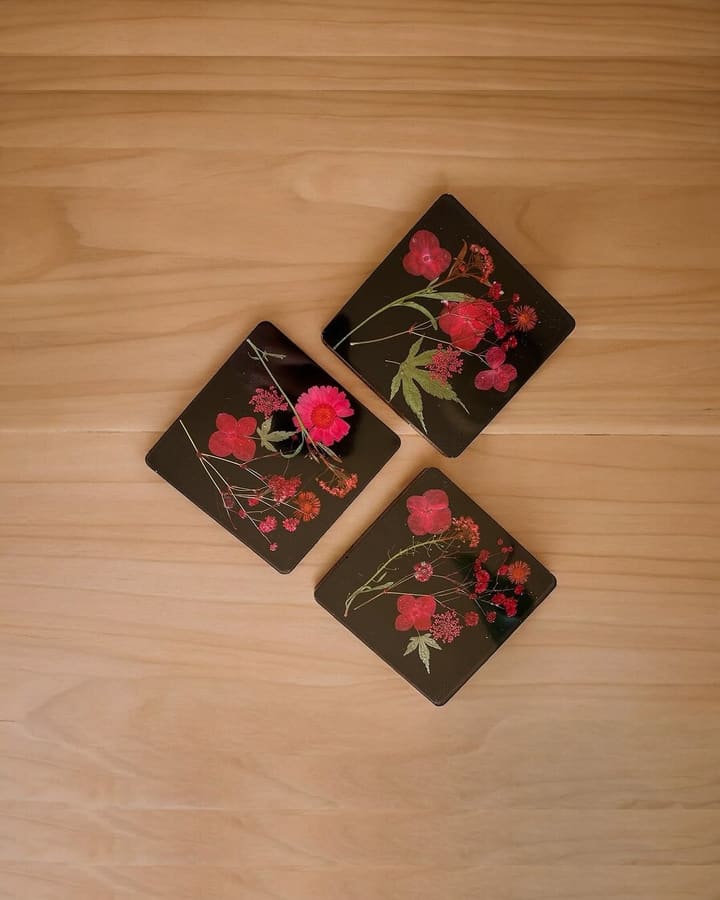 Three black square resin coasters with pressed red and pink flowers on a plain wood surface.