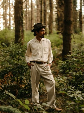 Octavio in a dark felt hat and a pale linen shirt and trousers, hands in his pockets, standing among ferns in a redwood forest.