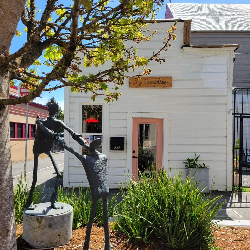 The front of La Ciendita on a sunny day: a white clapboard building with a carved wood La Ciendita sign, a pink door, a 655 mailbox and a bronze sculpture of two figures out front.