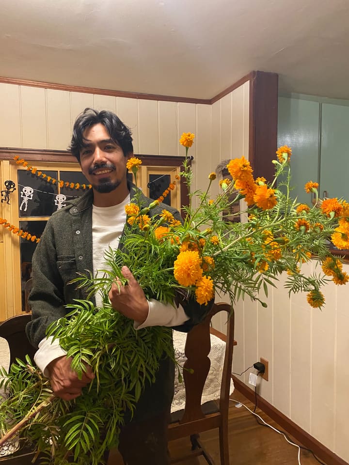 Octavio, smiling, holds an armful of orange cempasúchil in a room hung with marigold garlands and paper skeletons.