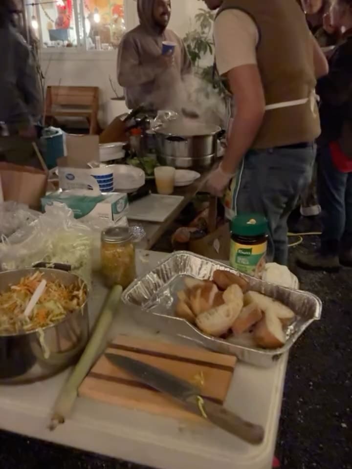 People cooking together at a night community dinner: steam rising from a big pot, bread on a tray, and several people gathered around the table.