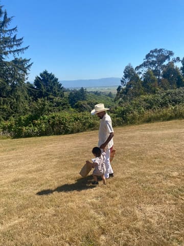 Octavio in a white hat walks a sunny field with a toddler.