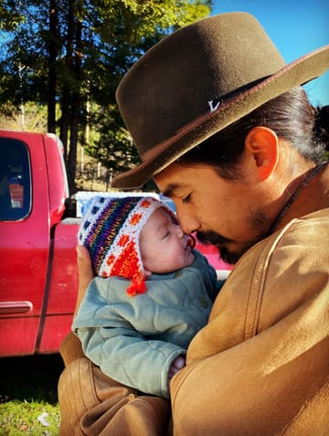 Octavio in a felt hat kisses a sleeping newborn in a bright knitted cap, a red truck and tall trees behind them.