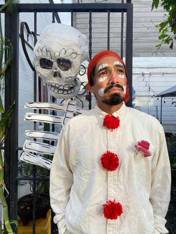 Octavio in white clown face paint and a red bandana, next to a papier-mâché skeleton on the courtyard’s iron gate.