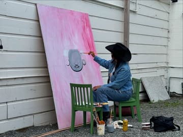 A painter in a black cowboy hat and denim jacket paints a face on a big pink panel in the gravel courtyard.