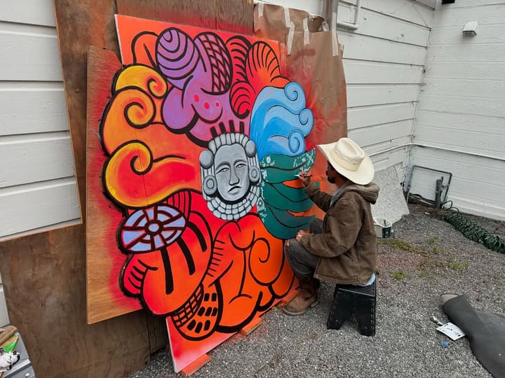 A painter in a straw cowboy hat and a brown work jacket paints the Nemiliztli panel in the gravel courtyard: a gray stone face ringed with bright orange, purple and blue swirls.