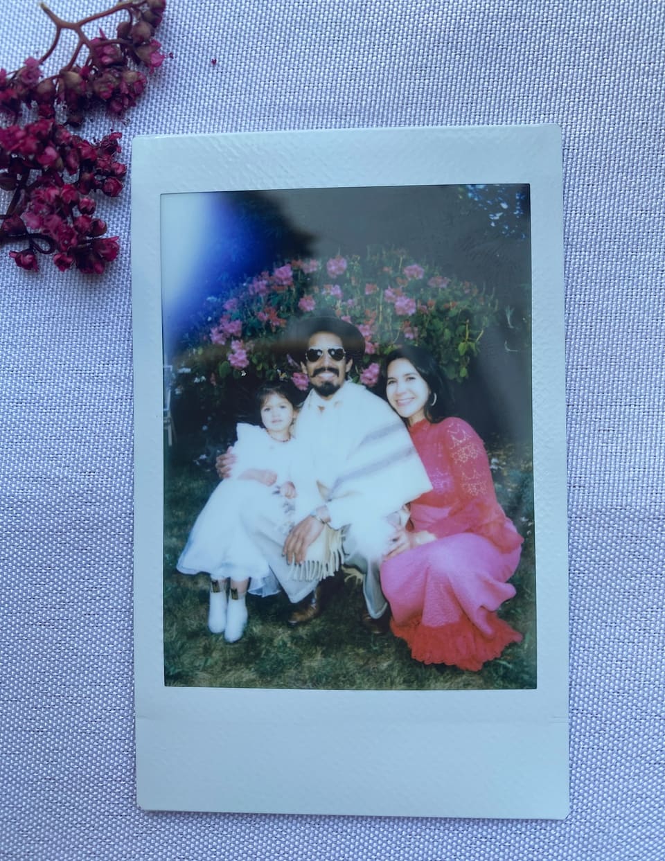 An instant photo of co-owners Octavio Acosta and Jessica Vanessa Olivera with their daughter: Octavio in a hat and sunglasses, Vanessa in a pink lace dress, their little girl in white, in front of a flowering bush.