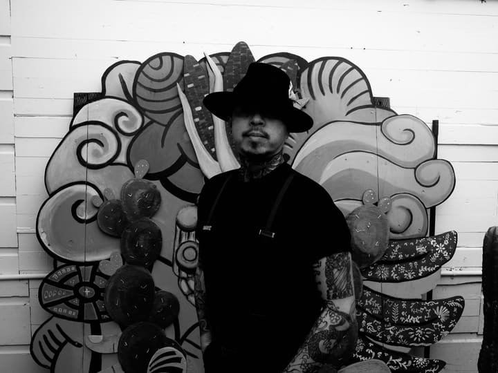 Black and white portrait of a man in a black brimmed hat and suspenders, tattoos on his arms, in front of the painted wood Nemiliztli installation.