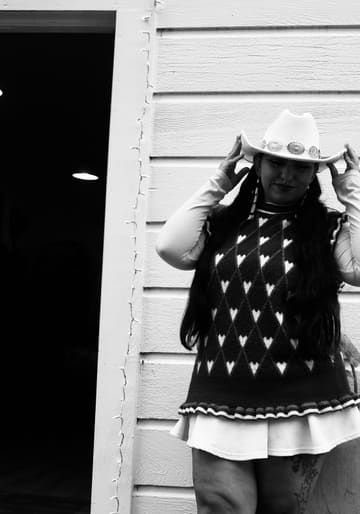 Black and white portrait of a woman in a white cowboy hat and a heart-patterned sweater vest, beside the shop door.