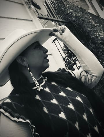Black and white profile of a woman tipping a white cowboy hat, long shell and bead earrings swinging.