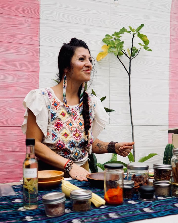 A smiling woman in an embroidered blouse and long beaded earrings stands at a table of herbs and jars, in front of the pink and white courtyard wall.