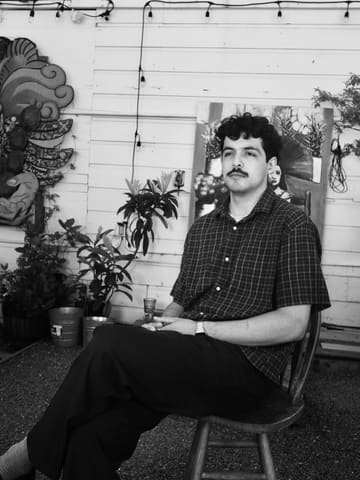 Black and white portrait of a young man with a mustache, seated on a wooden chair in the courtyard among plants and string lights.