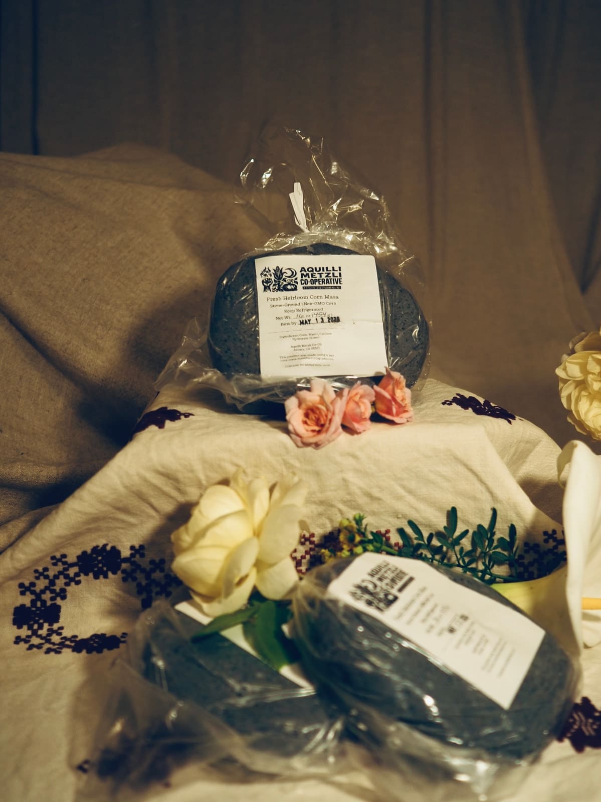 Three bags of fresh blue corn masa from Aquilli Metzli Co-operative on an embroidered cloth, one standing up and two lying in front, with pink roses, white roses and a calla lily.