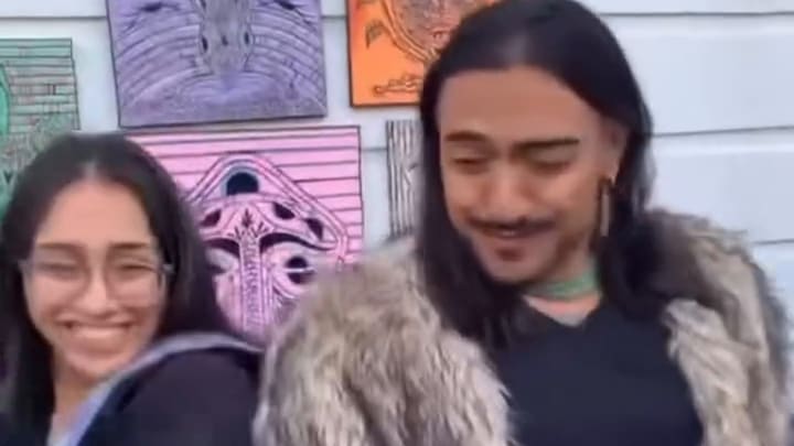 A woman in glasses and a man in a fur coat laugh together at an art pop-up, colorful prints hung on the courtyard wall behind them.