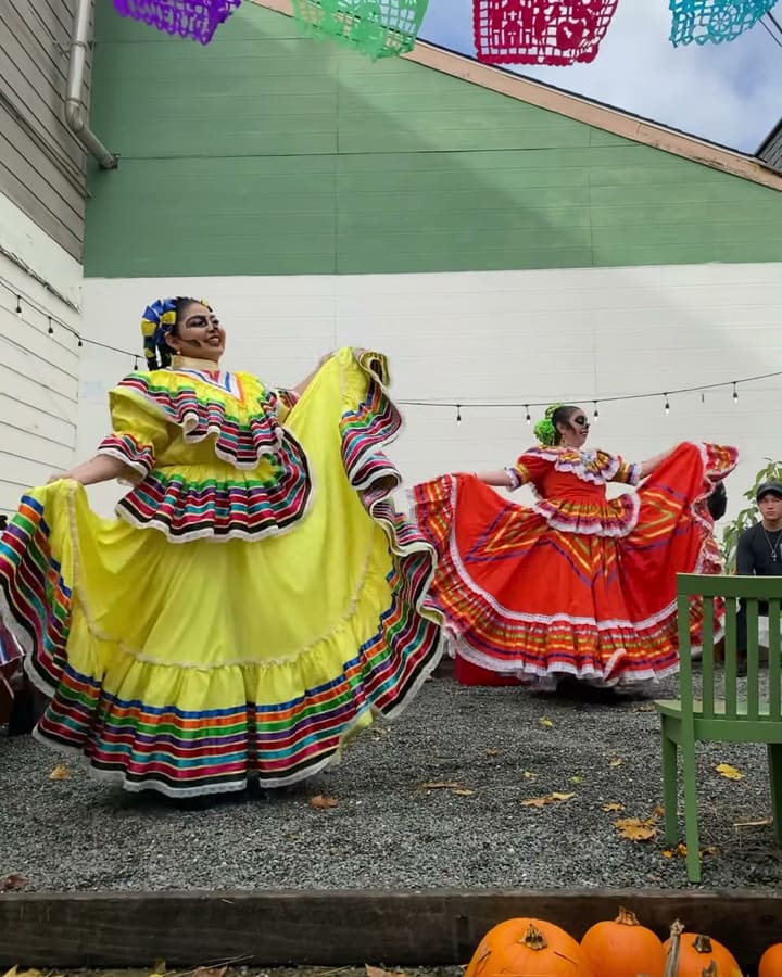 A smiling folklórico dancer in a yellow dress and calavera face paint holds her striped skirt out wide in the courtyard on Día de los Muertos, with a dancer in red behind her under papel picado.