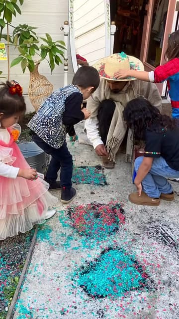 Kids and a grown-up in a straw hat crouch over a tapete of colored sawdust on Día de los Muertos.
