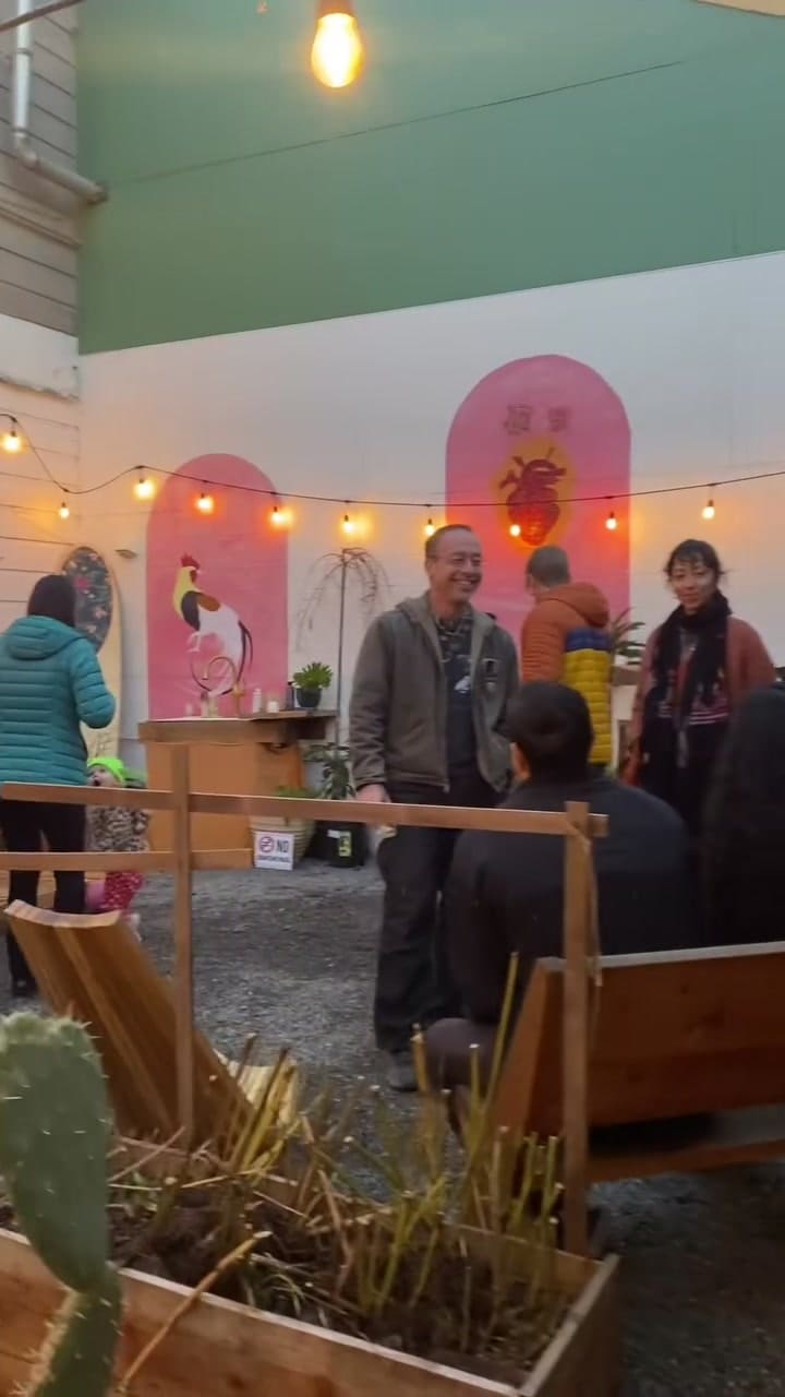 Friends gather in the courtyard at dusk for the Posada, string lights on and pink arch murals behind them.